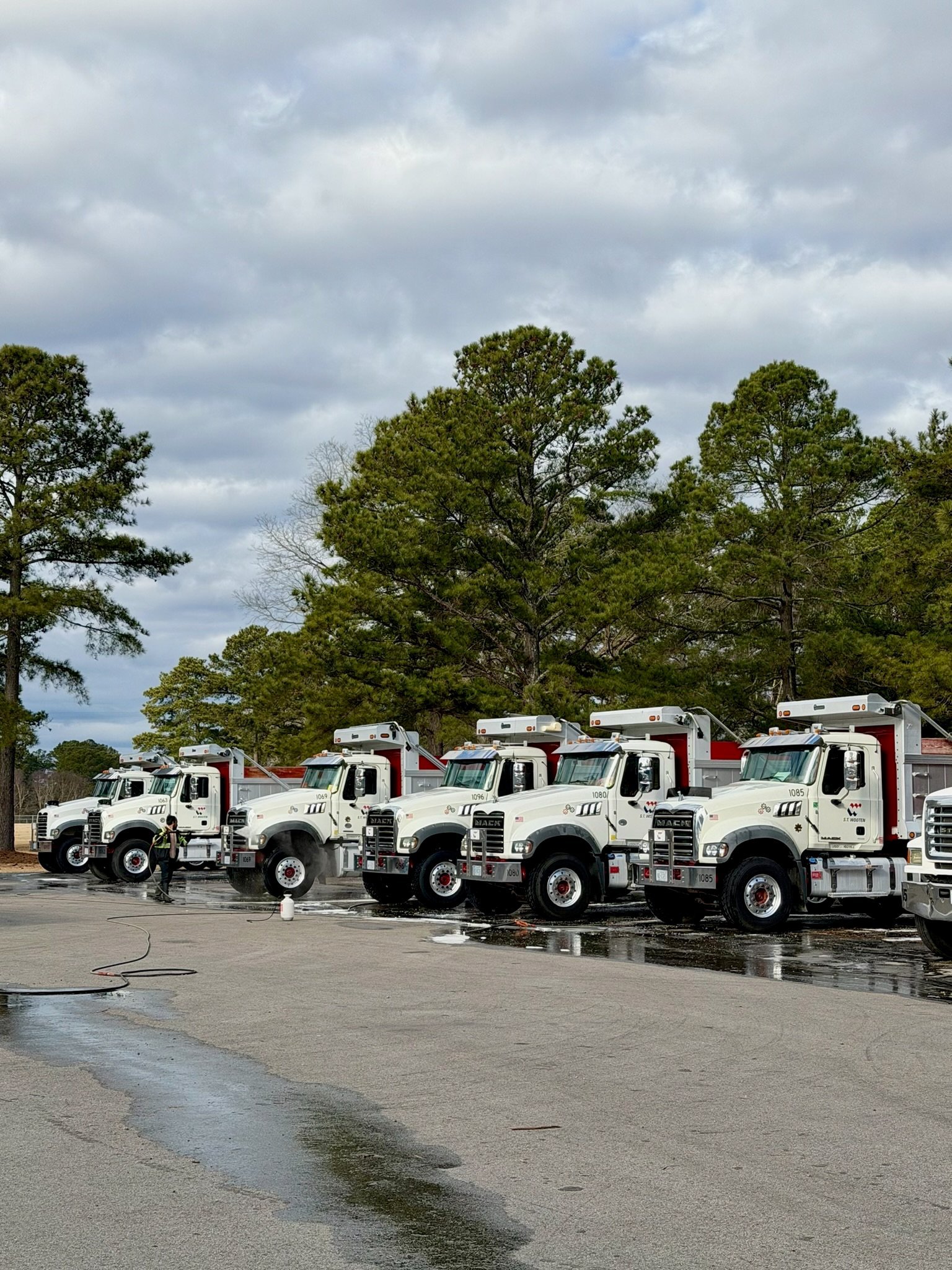 Row of white garbage trucks parked in a line on wet pavement with green trees and cloudy sky in background
