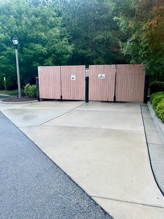 Wooden trash enclosure with four brown paneled doors on a concrete pad beside a tree-lined area and street lamp
