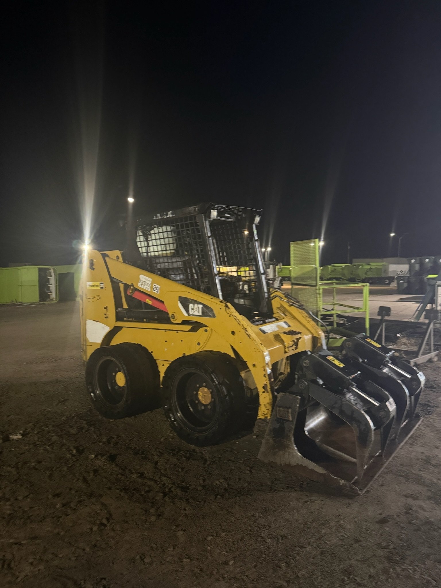 Yellow skid steer loader with bucket attachment parked at night in a construction yard with floodlights